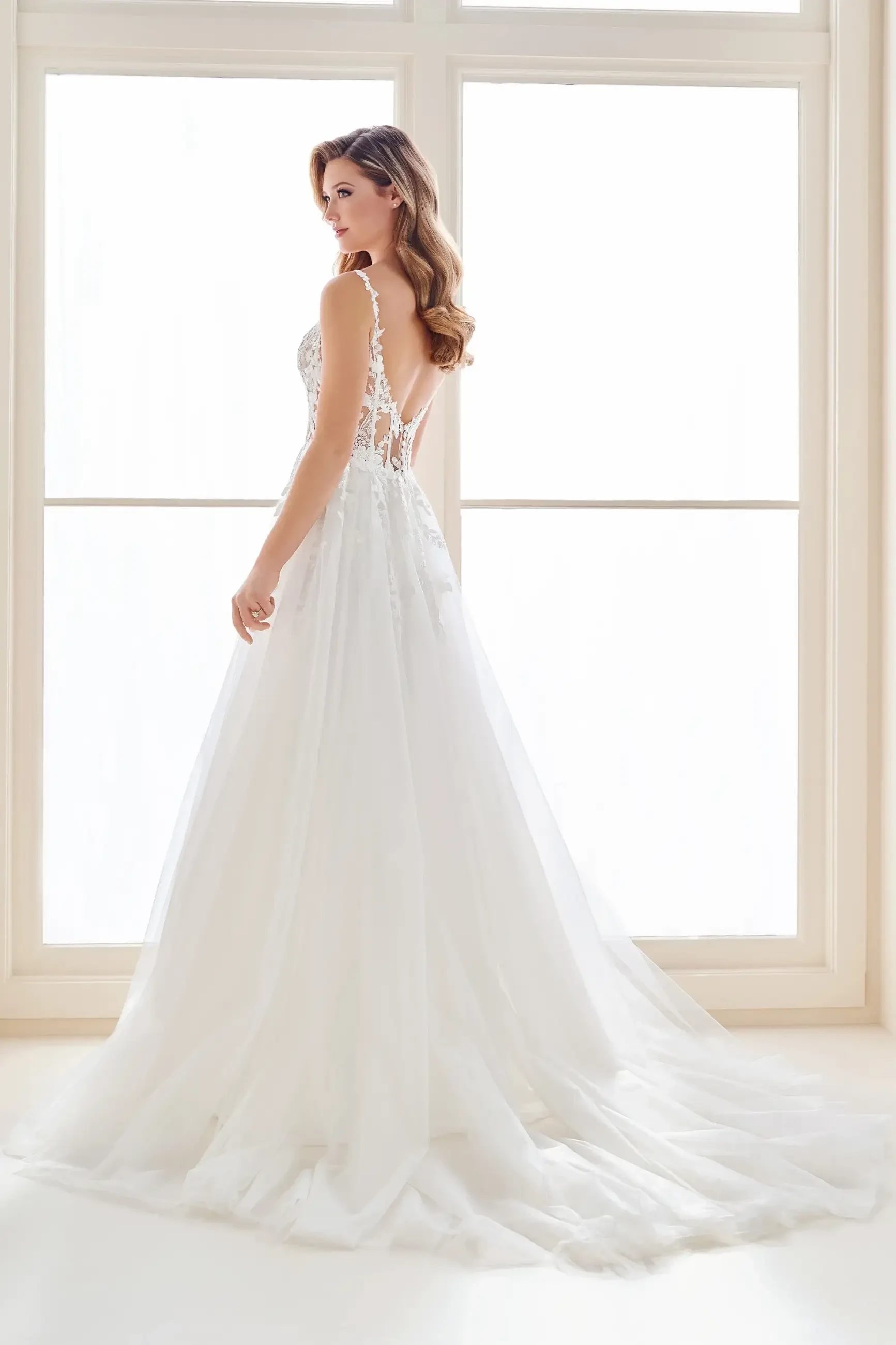 Model is wearing an A-line gown with a semi-sheer corseted bodice, deep plunge neckline, thigh split, and dramatic tulle train.