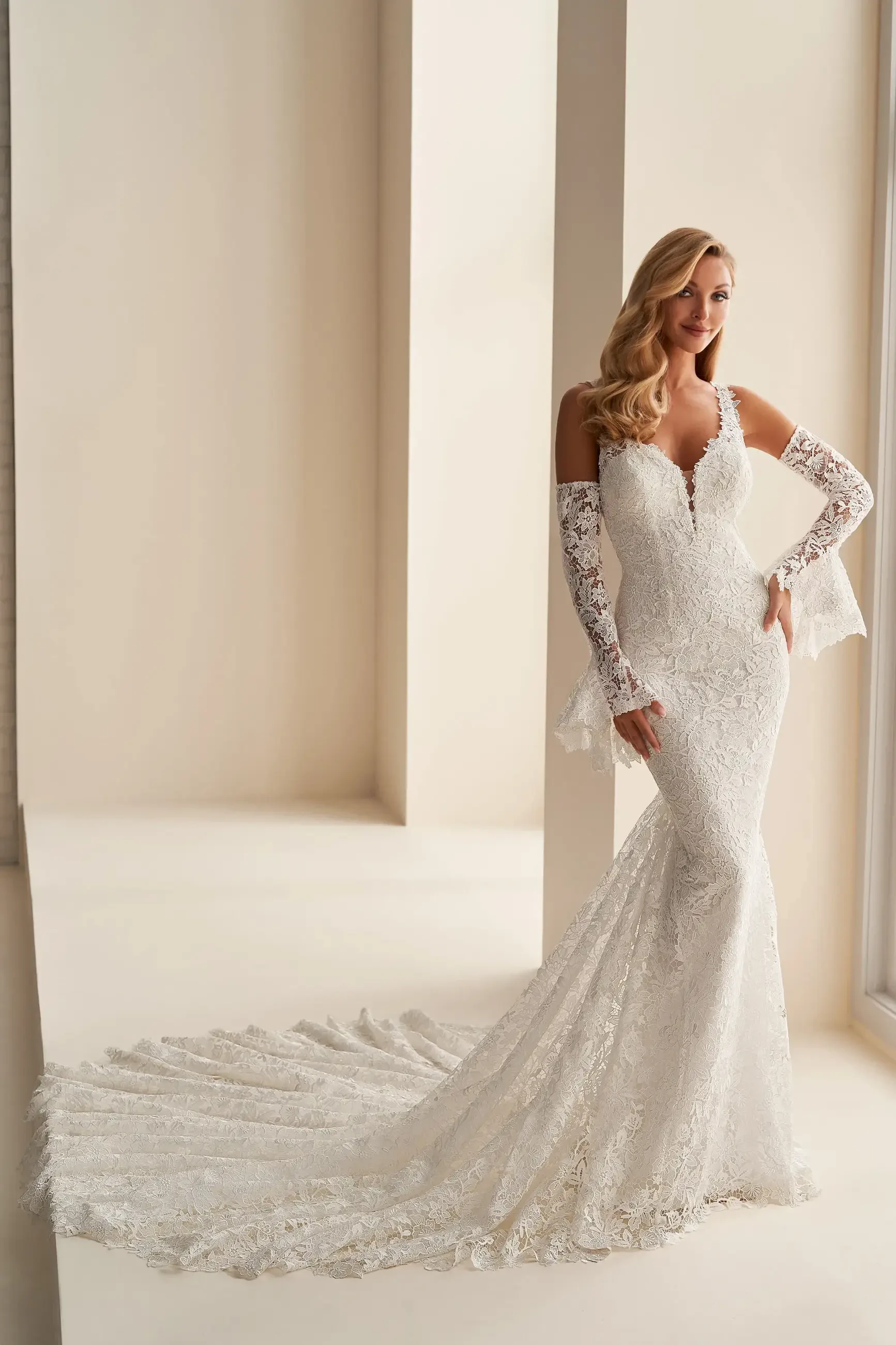 A model is posing in a beautiful white lace wedding dress with a fitted silhouette, long sleeves, and an intricate train. The background features soft, neutral tones, enhancing the elegance of the dress.