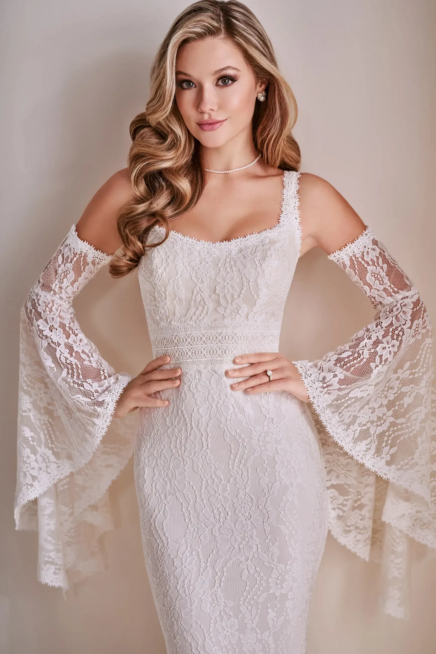 Model is wearing an allover lace mermaid gown with a scoop neckline, long lace train, and optional lace sleeves.
