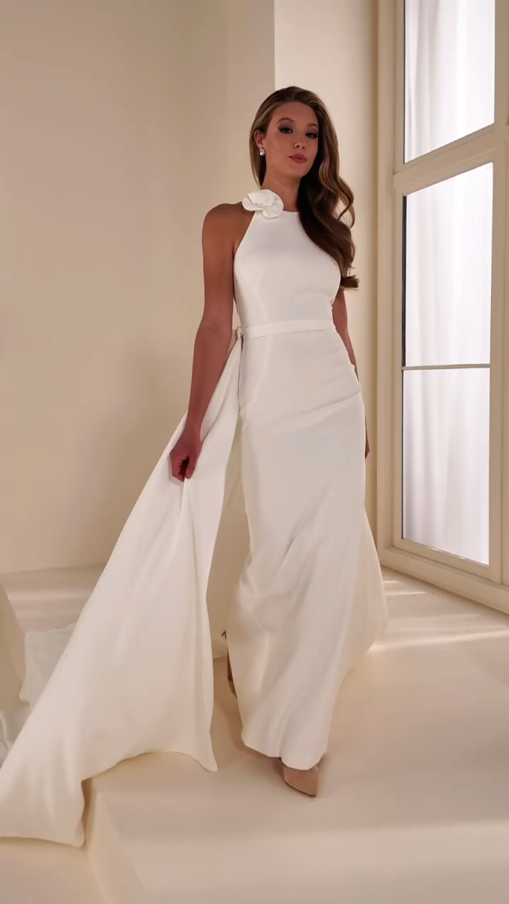 Model is wearing a sheath wedding dress in ivory with a high neckline, 3D flower accent, and detachable train.