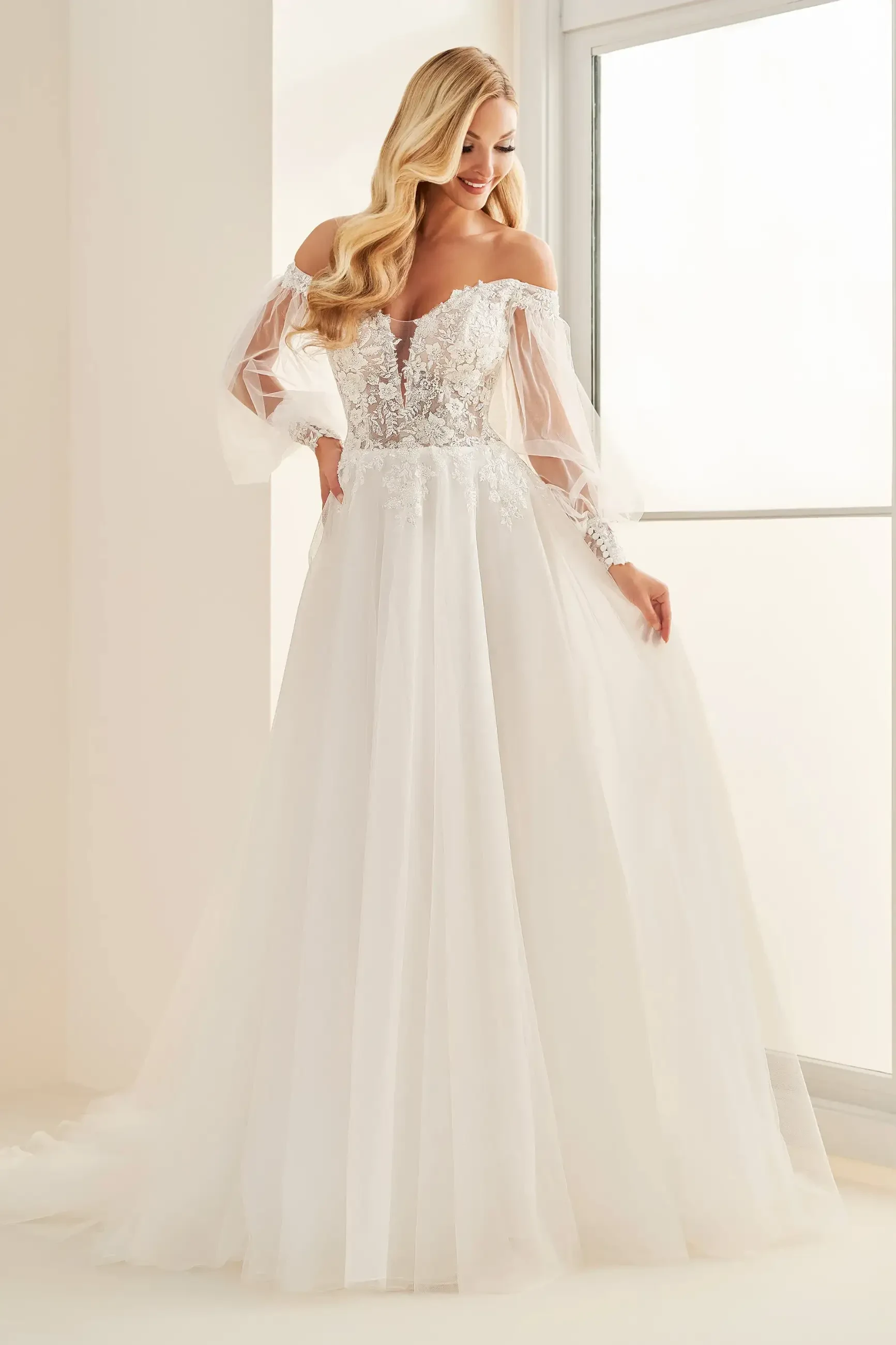 Model is wearing a strapless sweetheart ball gown with sequin lace appliqués, a full tulle skirt, and detachable bishop sleeves.