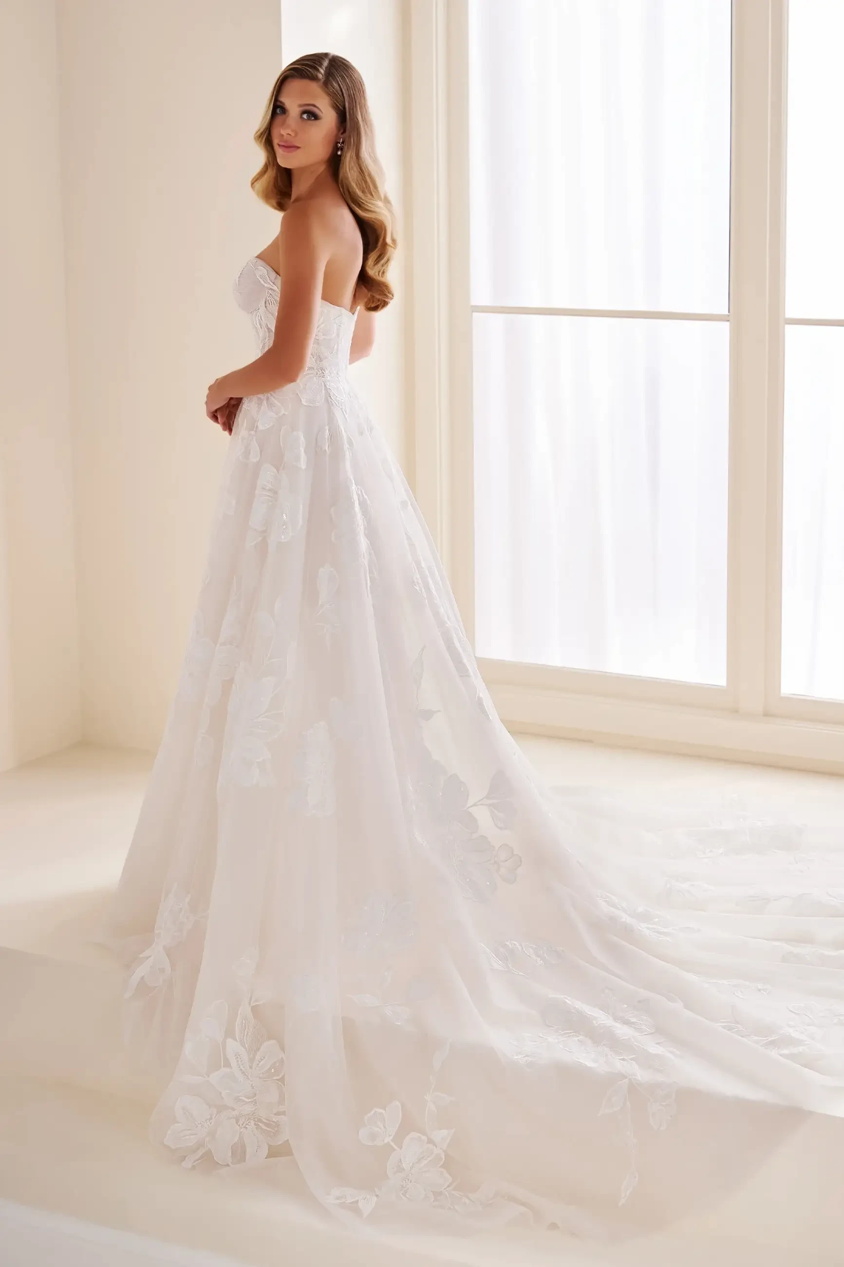 Model is wearing a romantic A-line gown with a sweetheart neckline, Schiffli lace appliqué, shimmering sequins, and layered tulle.