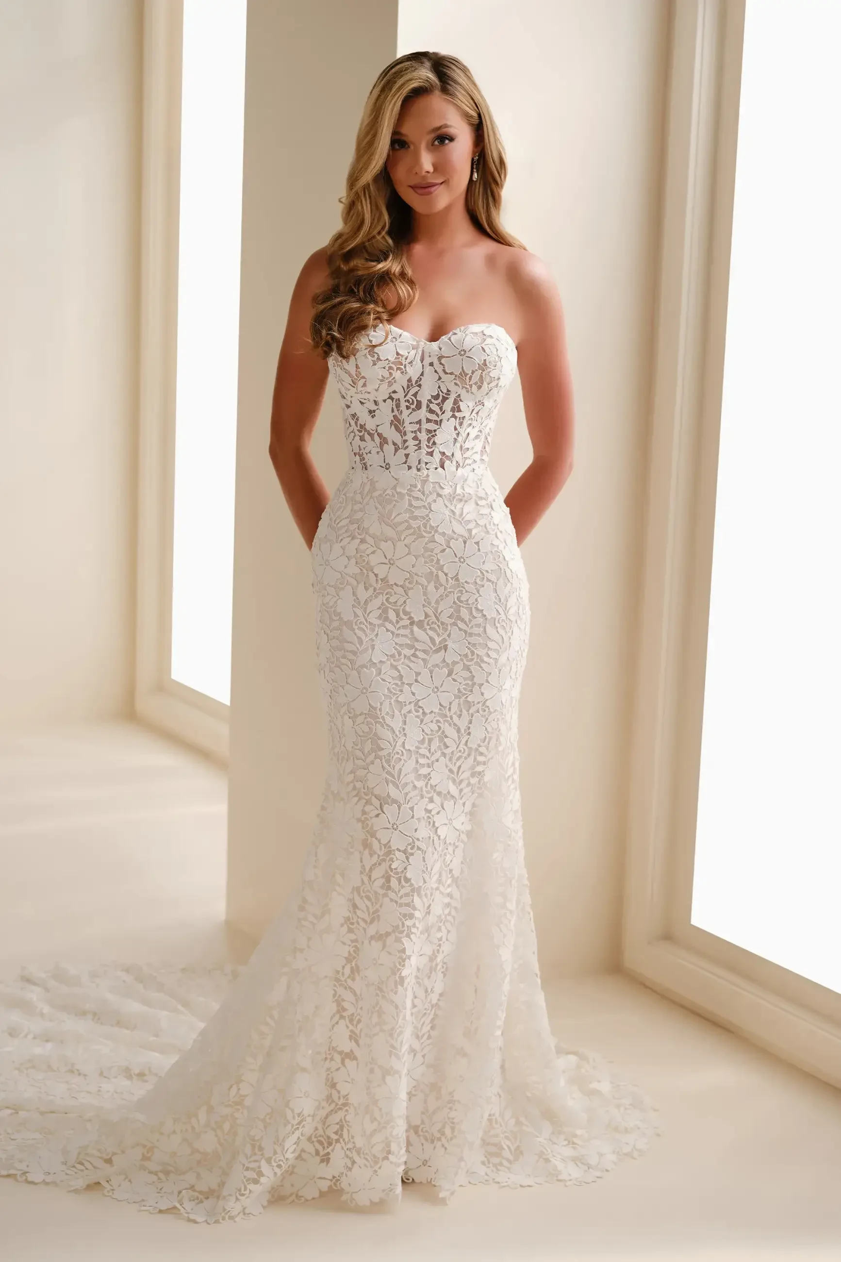 A woman wearing a strapless, lace wedding dress stands in front of large windows. The fitted gown has a floral pattern and a long, elegant train.