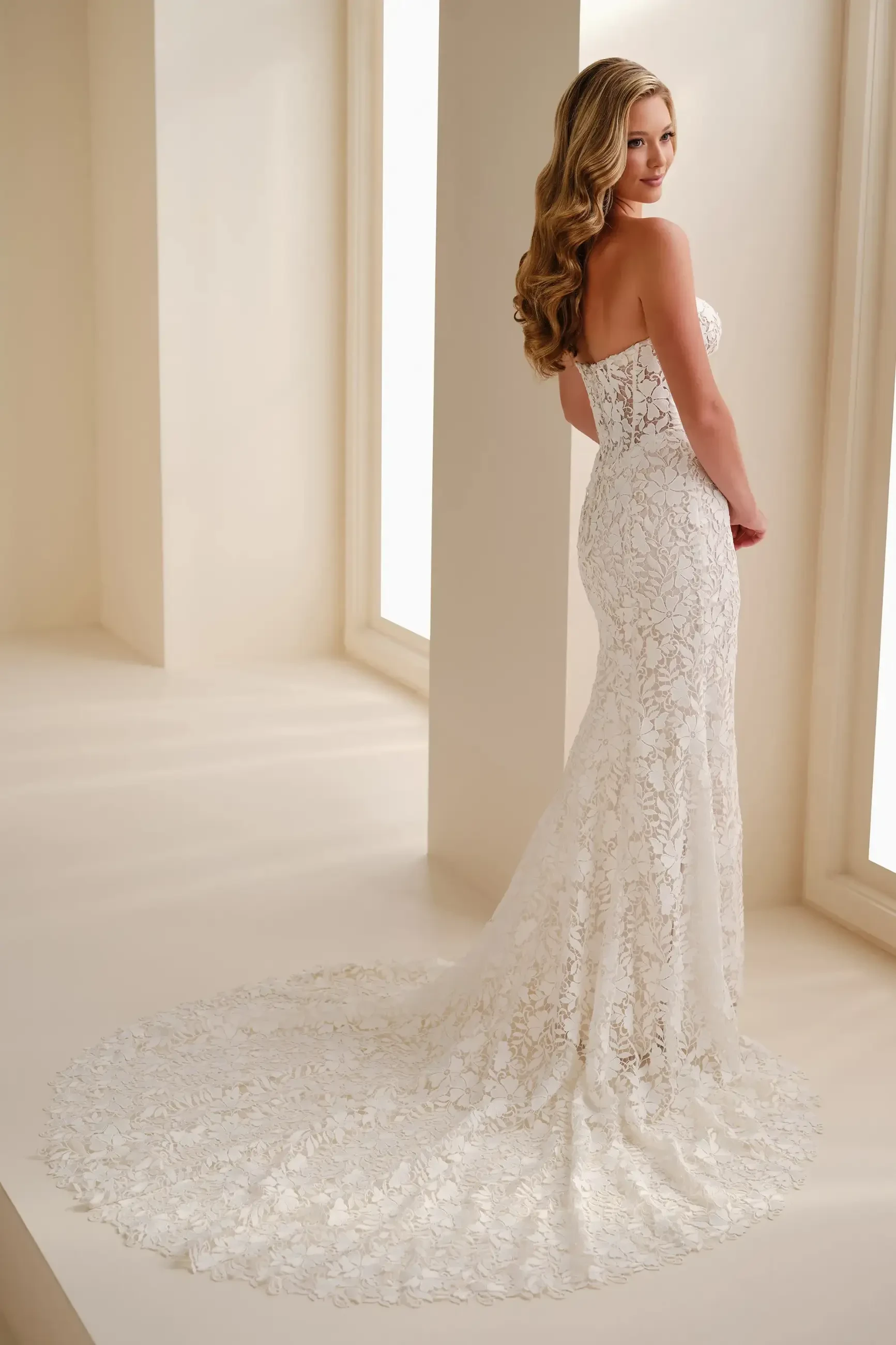 A woman wearing a strapless, lace wedding dress stands in front of large windows. The fitted gown has a floral pattern and a long, elegant train.