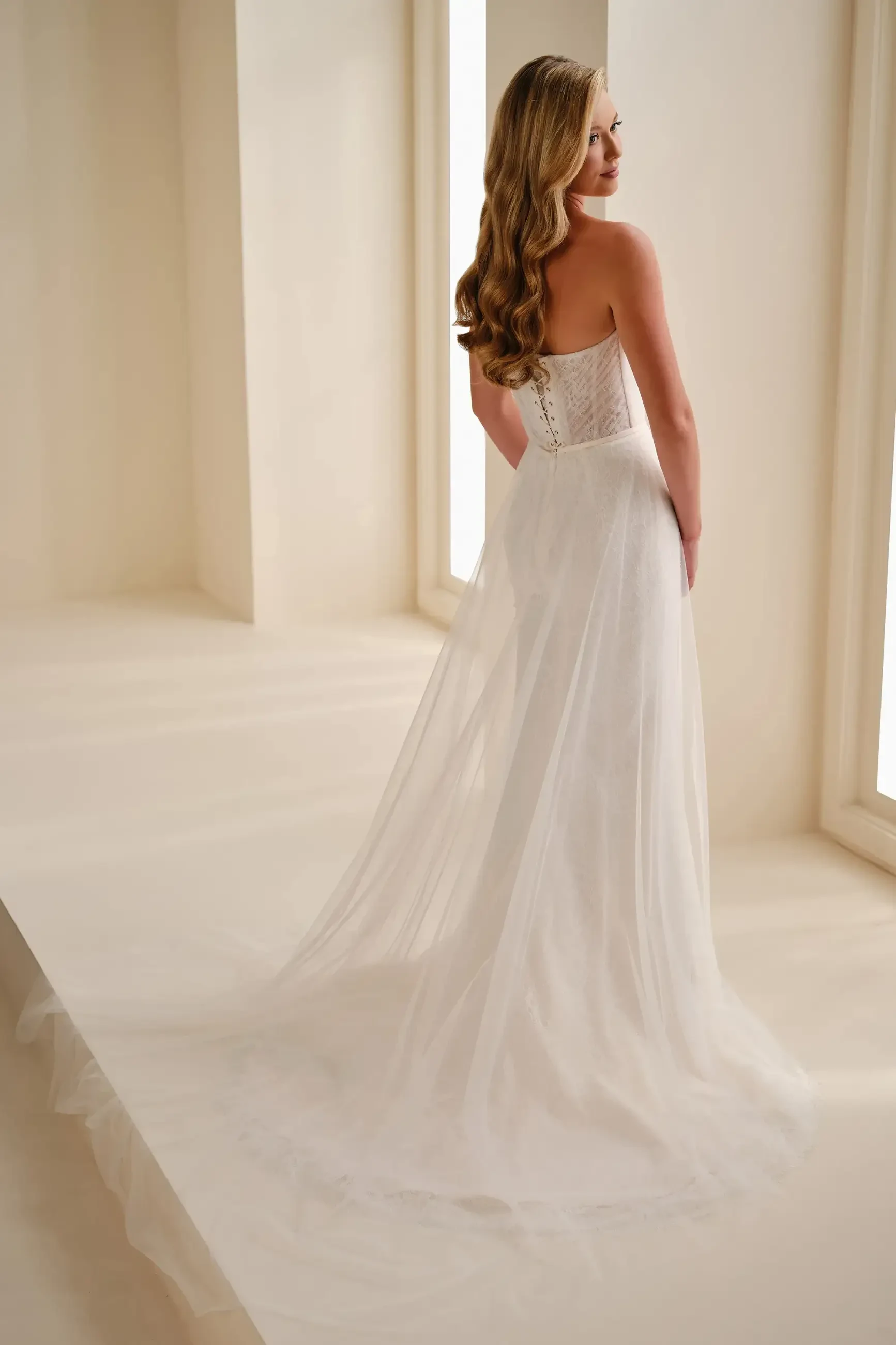 A woman in a flowing, strapless white wedding gown stands in a softly lit room with large windows. Her long hair cascades down her back, creating an elegant and serene atmosphere.