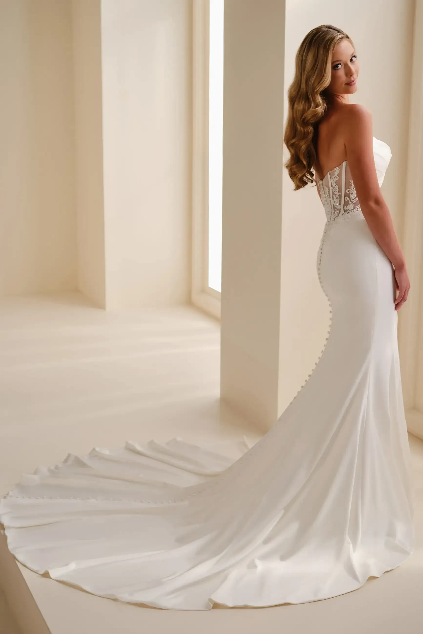 Elegant bride in a strapless white gown with lace back and long train, standing near a window in a softly lit room, exuding a serene, graceful mood.