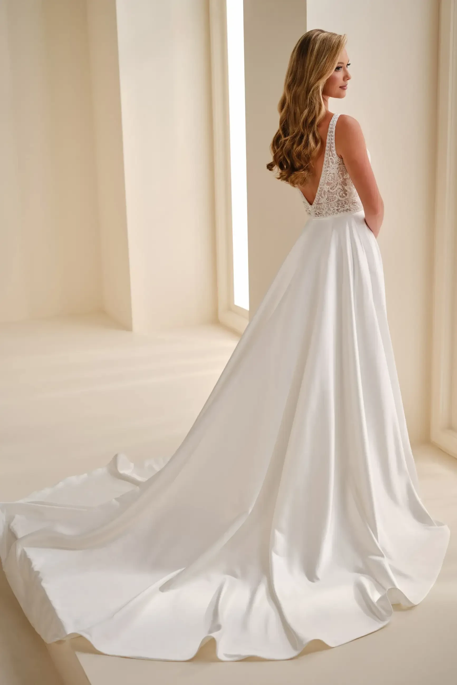A woman in a sleek white wedding dress with a long train and lace top stands in a minimalist room, gazing serenely out the window.