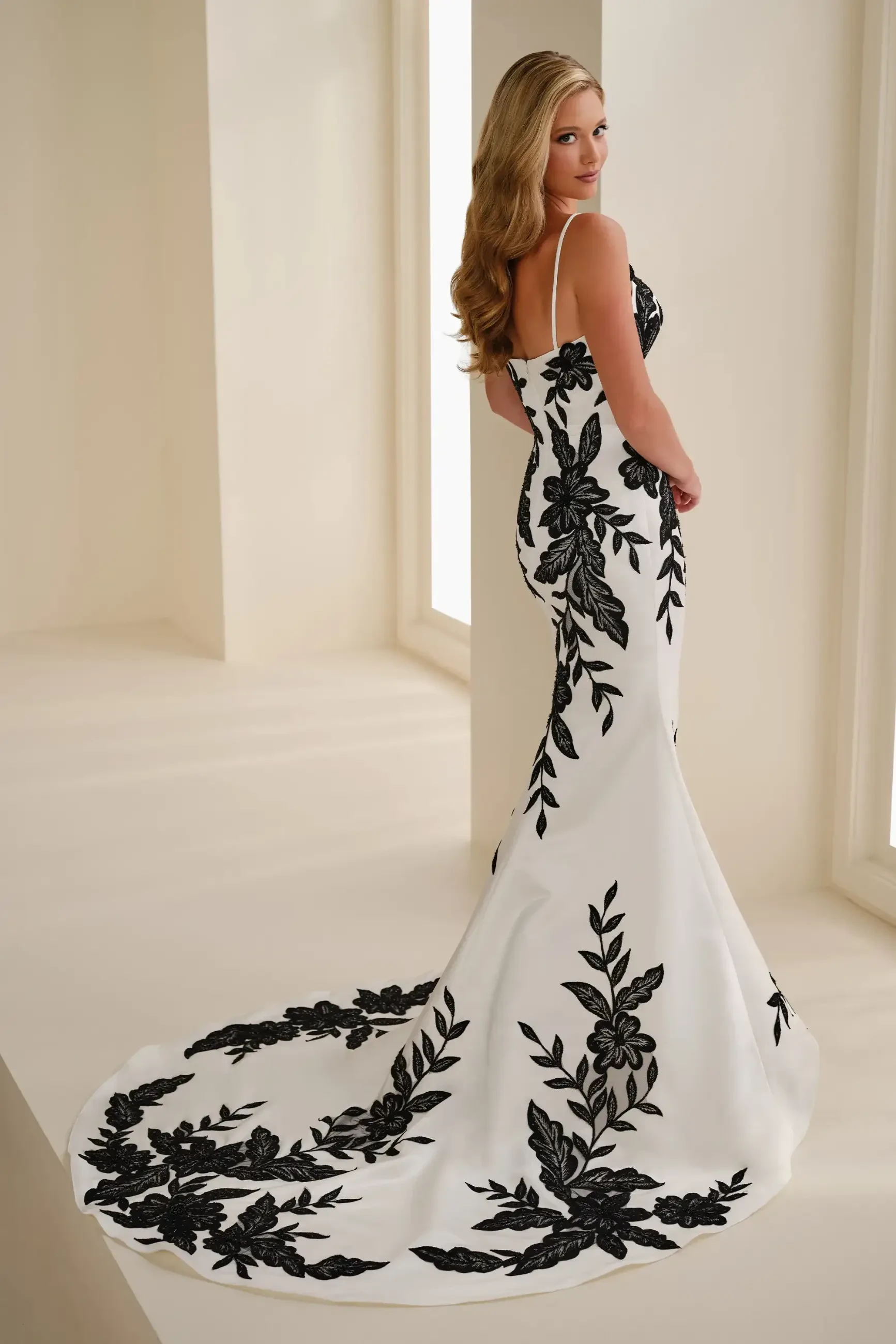 A woman in a white, floor-length wedding gown with floral embroidery stands in an elegant, softly lit room. Her expression is serene and confident.