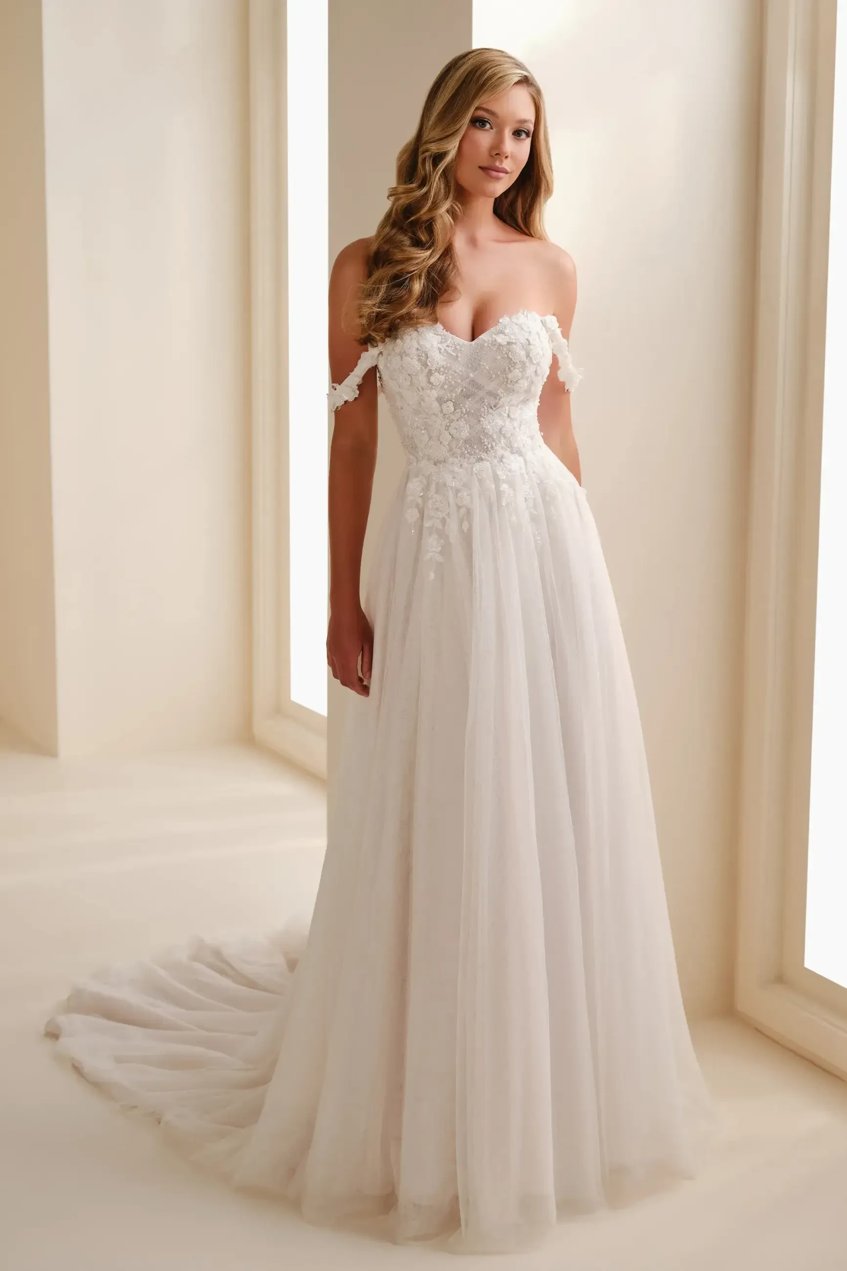 A woman in a strapless, lace-embellished white wedding gown stands elegantly in a softly lit room. The gown has a full skirt and a train.