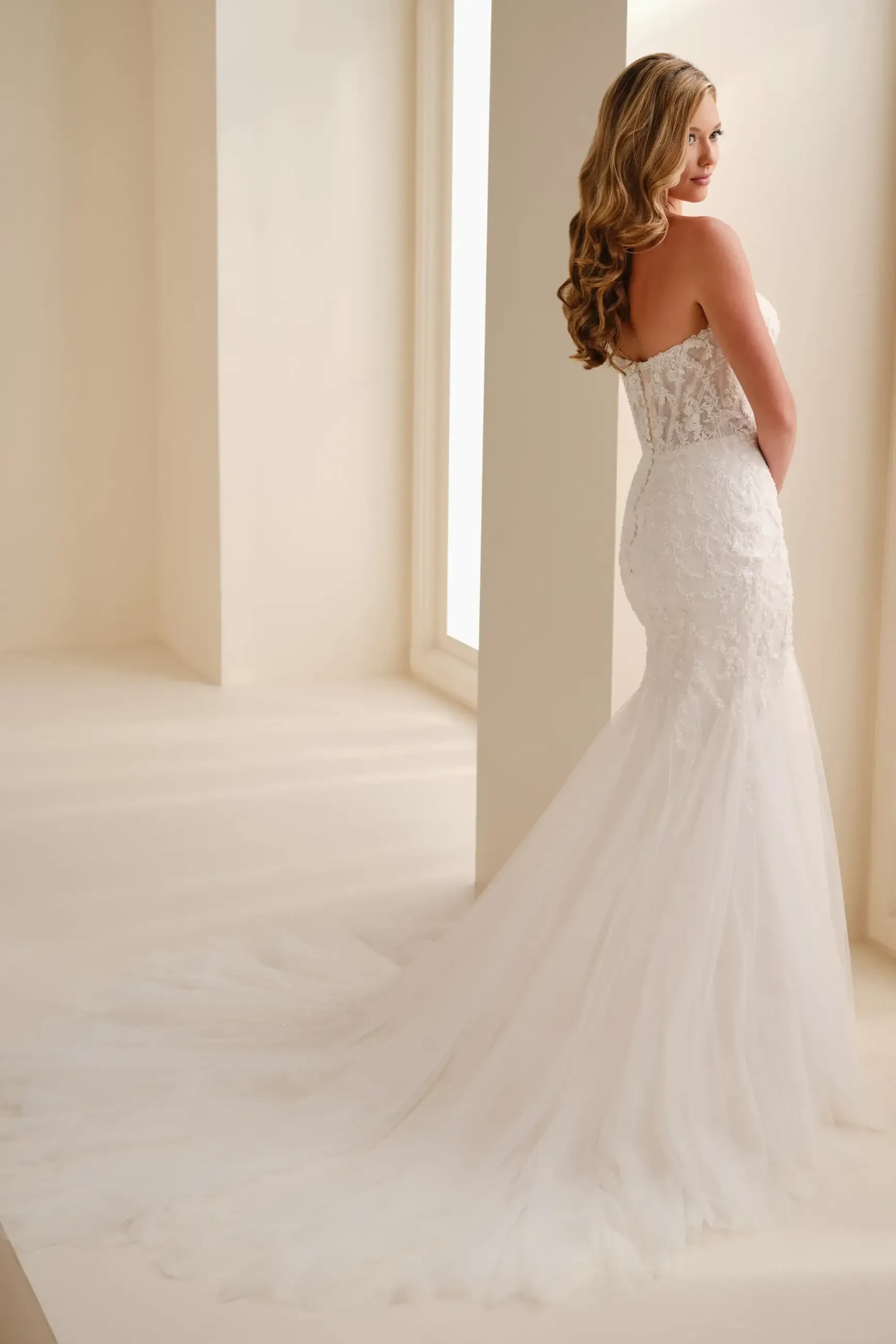 A woman in a strapless, lace wedding gown with a fitted bodice and flowing tulle mermaid skirt stands gracefully in a softly lit room, exuding elegance.